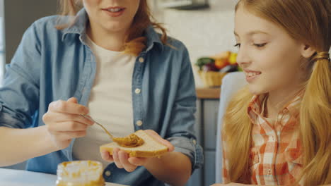 Young-attractive-mother-spreading-a-peanut-butter-on-the-bread-and-talking-with-her-teen-daughter-while-they-sitting-at-the-table-at-the-kitchen.-Close-up.-Inside